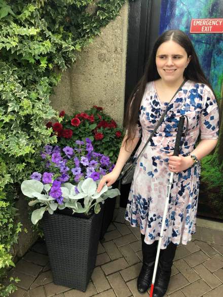Meagan, wearing a colourful summer dress and tall black boots, smiles as she touches soft, fuzzy leaves. Purple petunias are visible in the background.